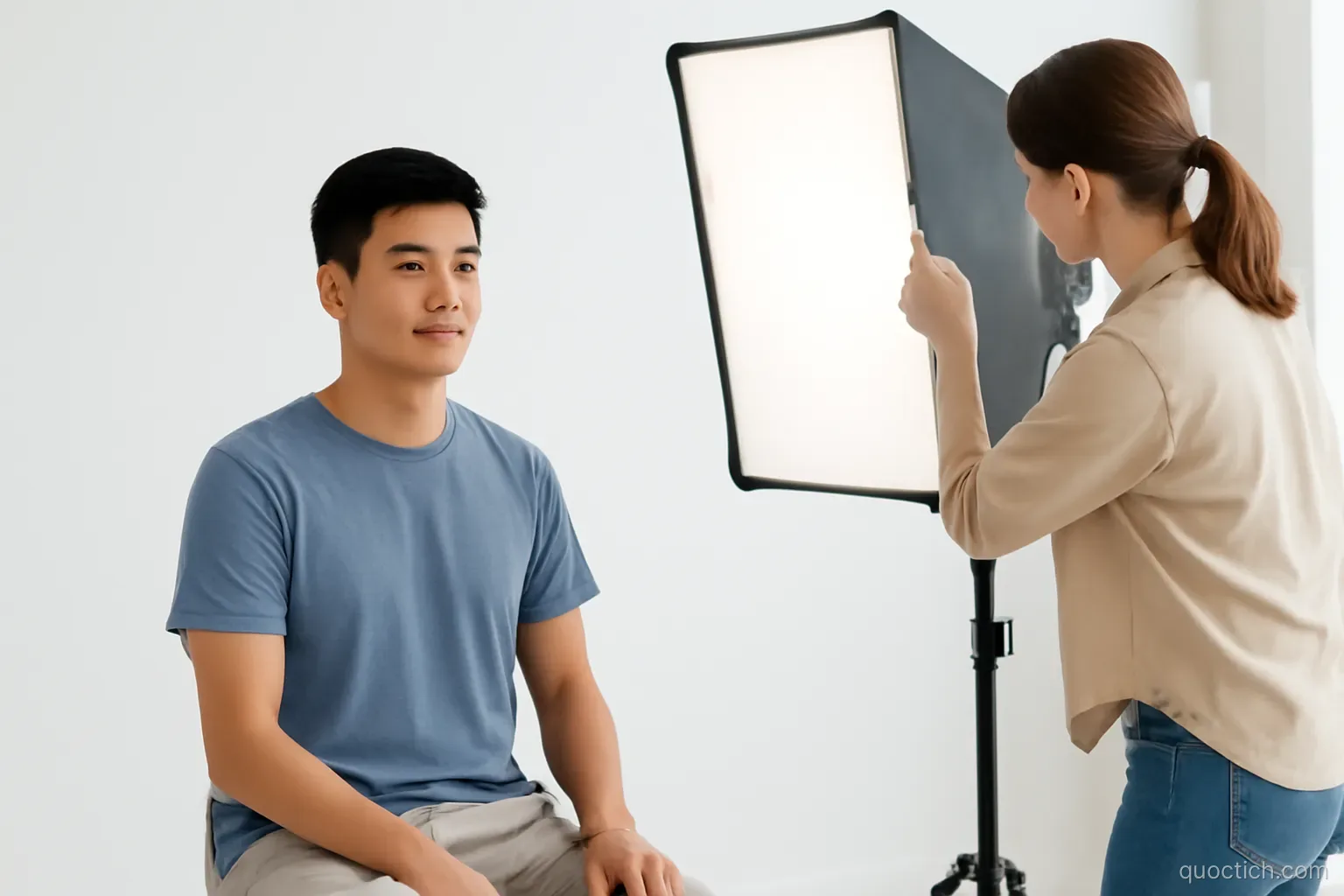 Young Vietnamese man sitting upright before a plain white backdrop in a bright modern photo studio while a female photographer adjusts a large softbox light, medium shot, clean white tones